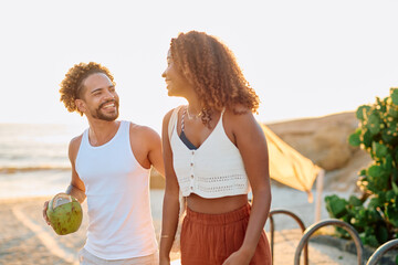 Happy couple smiling and walking together on a tropical beach during golden hour, man holding a...