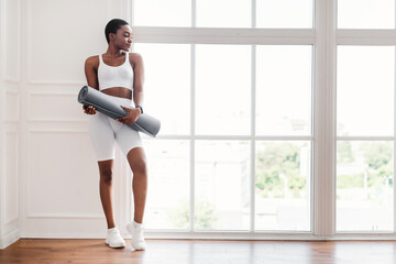 Obraz premium Ready For Training. Portrait of African American fit woman going to gym, holding and carrying roll mat standing by window in fitness studio or living room, looking at free empty space, panorama