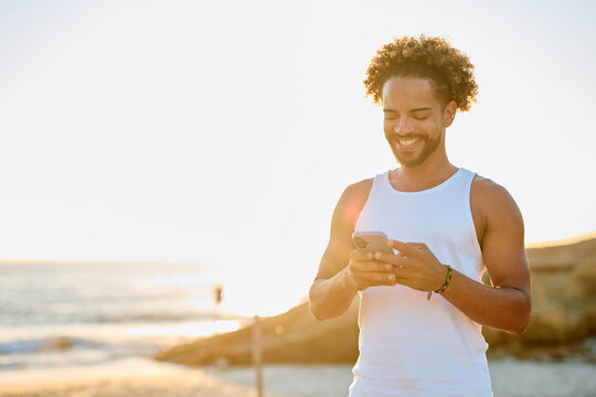 Young man smiling, wearing a tank top, interacting with a smartphone at a beach during a vibrant sunset - Powered by Adobe