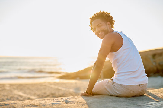 Young man with curly hair smiling and looking back at the viewer while sitting on a wall at a beach during sunset