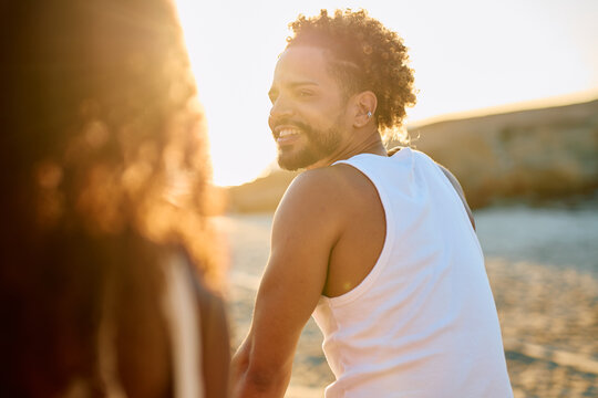 Smiling man with curly hair looking back at companion on beach during scenic sunset, experiencing happiness and friendship