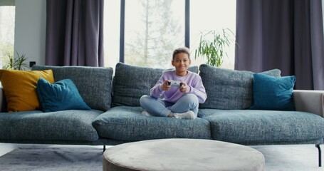 Obraz premium Little girl playing video game with joysticks sitting on the sofa at home