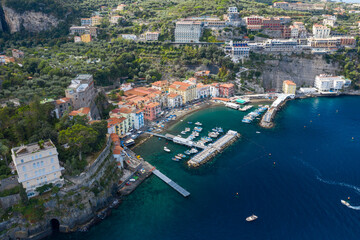 Obraz premium High-angle panorama of Sorrento, Italy, with vibrant houses clustered around a small marina and dramatic cliffs rising behind. The scene features turquoise Mediterranean water, lush greenery, and a