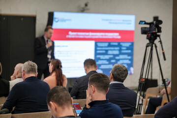 Audience seated in modern conference room watching speaker on stage presenting slides. Camera records event while attendees focus on content and notes.