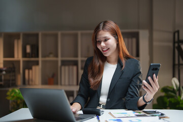 Young asian businesswoman in an office working late, smiling while using a laptop and a smartphone, demonstrating productivity and successful communication in a modern workspace