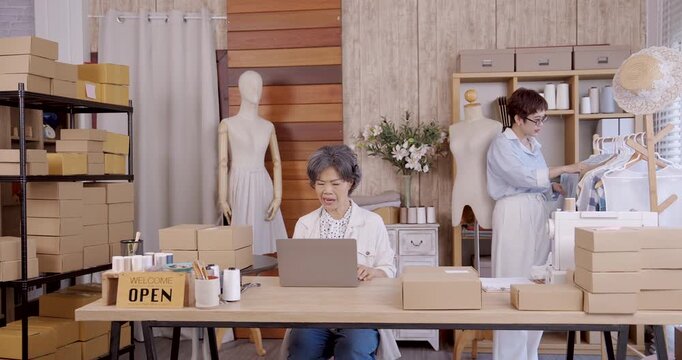Senior asian mother and adult daughter giving high five smiling at desk with laptop and packages, teamwork celebrating online shop achievement for small family boutique fashion business e commerce