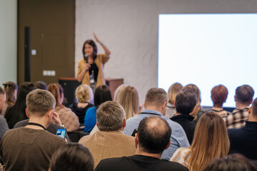 Speaker presents to a busy audience during a conference. Attentive listeners focus on the speaker while a large screen glows in the background.