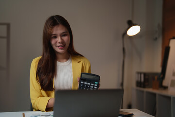 Young Asian business woman presenting financial data using a calculator during a virtual meeting on her laptop, working on accounting and budget management in her office