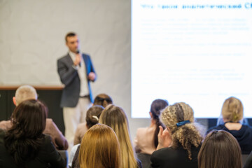 Presenter speaks to attentive audience in bright conference room. Large projection screen displays text. Attendees listen, take notes, and engage with talk in professional setting.