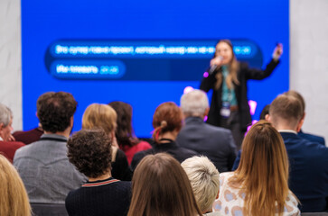Speaker presents to attentive audience at conference with bright blue backdrop. Audience listens closely and engages.