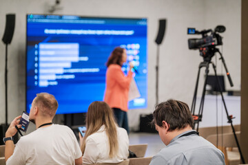 Conference room scene with speaker on stage delivering content to audience while camera records. Large blue screen shows charts. Attendees watch smartphones and take notes.