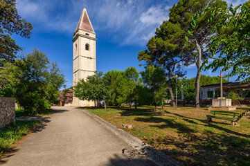 Stone bell tower rising above pine trees on the Croatian island of Silba, along a quiet path under blue sky. Mediterranean village atmosphere, summer travel, history, peace, and island serenity calm