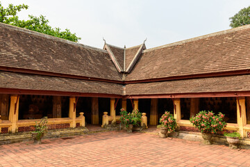 Naklejka premium Symmetrical view of the historic gallery at Wat Si Saket temple in Vientiane, Laos, featuring a traditional sloped tiled roof, yellow columns, and potted flowering plants. The brick courtyard and