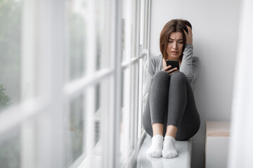 Confused shocked sad depressed european millennial lady sits on windowsill and reading message on smartphone, receiving bad news. Breakup, stress, mental problems and gadget addiction, copy space