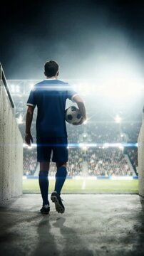 Silhouette of a soccer player holding a ball in a dark tunnel walking towards a bright stadium. Football athlete entering the arena pitch for a night match. Vertical video for social media