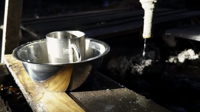 Shiny metal bowl and mug near old leaking pile in dugout. Tourist dishware stands on wooden shelf against dripping water in frontline blindage
