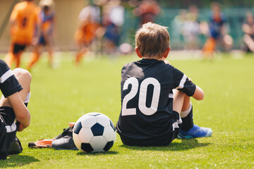 Young Soccer Player Resting During Match. Child Sitting on Grass With Football, Youth Sports Moment Outdoors