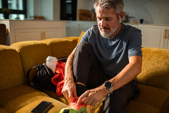 Mature man tying running shoes on living room sofa