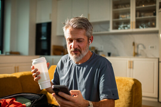Middle aged man with earbuds using smartphone and drinking protein shake in home kitchen