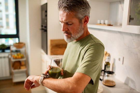 Middle-aged man checking smartwatch while drinking green smoothie in modern kitchen