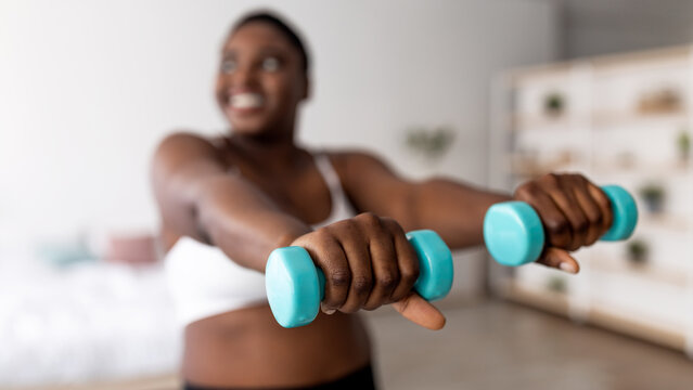 Domestic strength training. Plump black woman doing exercises with dumbbells, losing weight, strengthening her body at home. Overweight Afro lady working out biceps muscles, selective focus. Panorama