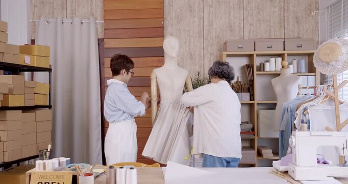 Senior asian mother adjusting fabric on mannequin while adult daughter discussing design beside, teamwork creating handmade garment pattern for family boutique fashion small business online shop
