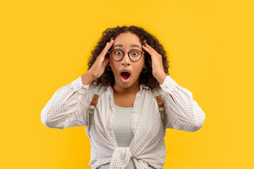 A woman stands with her hands on her head, eyes wide in surprise. She wears glasses and a casual...