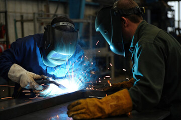Two industrial welders working together during metal welding process in factory workshop.