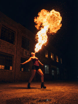 Female fire breather performing dramatic flame show at night in urban industrial setting.