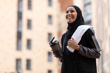 Glad young muslim lady in hijab and sportswear with phone on shoulder with cocktail cup and towel...