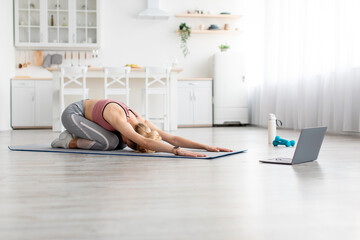 European millennial blonde sportswoman doing body stretching and watching online workout lesson on laptop at home. Athlete practices yoga and exercises, online blog remotely during covid-19 quarantine