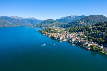 High-angle panorama of Orta San Giulio on the shores of Lake Orta in northern Italy, with deep blue water, clustered lakeside buildings, and forested hills rising in the background. The scene features