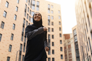 Fototapeta premium Portrait Of Young Muslim Woman In Modest Sportswear Jogging On City Streets, Low Angle View Of Sporty Islamic Lady In Hijab Running Near Urban Buildings, Enjoying Training Outdoors, Free Space