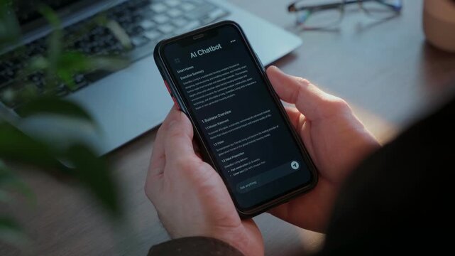 Close up of hands holding smartphone with AI chatbot app at workspace with laptop. Artificial intelligence assistant application helps improving business productivity. 