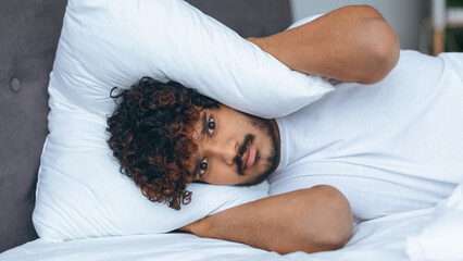 A man lies in bed with curly hair, covering his ears with a pillow, looking frustrated as morning...