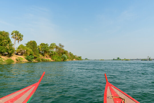 Two pointed red wooden boat prows frame a wide stretch of the Mekong River in the 4000 Islands region of southern Laos. Clear daylight reveals rippling blue water, tree-lined riverbanks, and scattered
