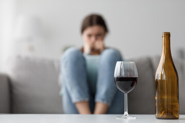Sad caucasian millennial female sits on sofa suffering from stress and troubles, focus on bottle and glass of wine in home interior. Alcoholism, drinking alone, depressed, addicted to alcohol at home