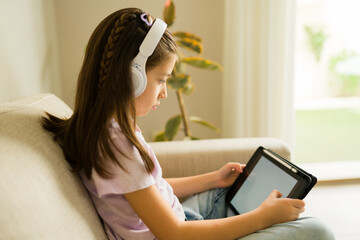 Little girl wearing headphones and holding digital tablet, focusing on e-learning and remote education at home