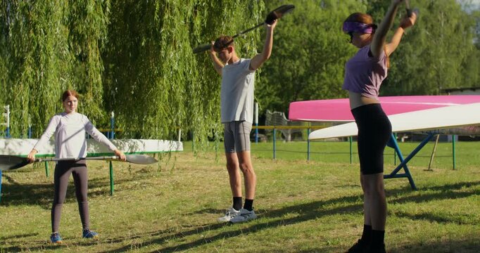Two girls and a guy hold oars in their hands and perform warm-up exercises. Male and female rowers do warm-up exercises before training.