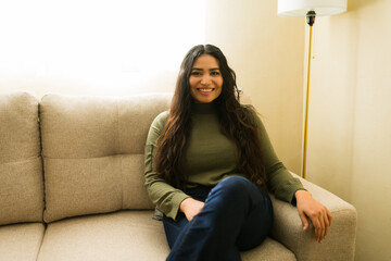 Obraz premium Latin woman relaxing on a sofa at home, smiling with long dark hair, enjoying comfortable domestic living