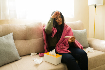 Woman feeling unwell at home, checking her temperature with a thermometer while suffering from fever and flu symptoms