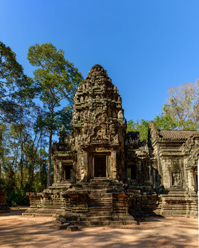 The main sandstone tower of Thommanon temple rises under a clear blue sky, surrounded by tall forest trees in Angkor, Cambodia. Crisp sunlight highlights the intricate carvings and weathered stonework
