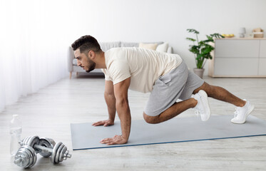 Fototapeta premium Active lifestyle during covid lockdown. Handsome young Arab guy in sportswear exercising on yoga mat, doing domestic fitness, working out his legs muscles at home, side view