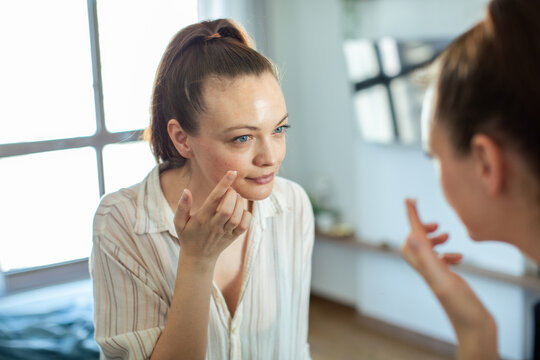 Young woman applying face cream in front of mirror at home