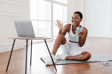 Excited athletic black female in sportswear waving to webcam making video conference with personal trainer or followers, sitting on the floor on yoga mat in front of laptop, holding bottle of water