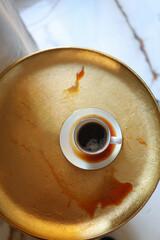 Coffee cup on saucer with spilled espresso on a gold textured surface near window light. Artistic composition about real life moments, mess, authenticity.