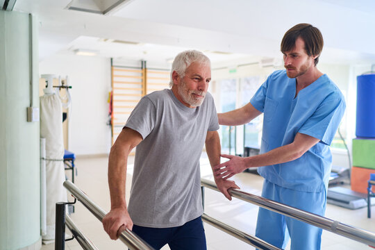 Senior Man Walking With Parallel Bars Assisted By Physiotherapist In Rehab Center For Injury Recovery And Mobility Training Indoors