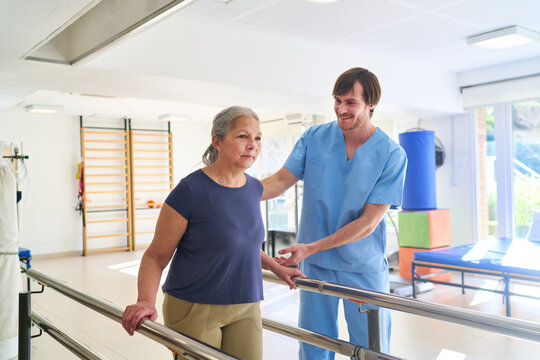 Senior Woman Walking with Physiotherapist Assistance on Parallel Bars for Rehabilitation in Rehab Center Indoor Therapy Session