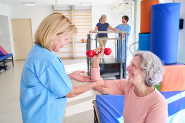 Senior Woman Lifting Dumbbell With Physiotherapist Assistance During Rehabilitation Exercise Indoors At Rehab Center For Recovery And Strength