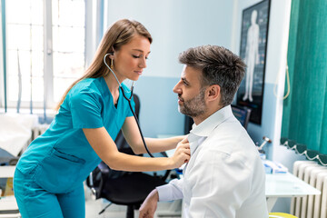 Obraz premium Female doctor in blue scrubs using stethoscope to listen to male patient's heart. Professional healthcare specialist performing chest auscultation during a clinical medical consultation.